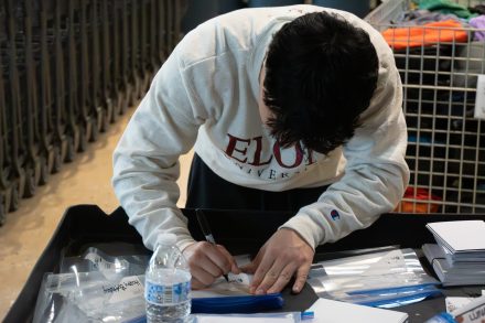 Vaughn Cultrara ’27 writes on a label while volunteering at Classroom Central. They are leaning over a table filled with supply bags, note cards, and a water bottle, wearing a white Elon University sweatshirt.