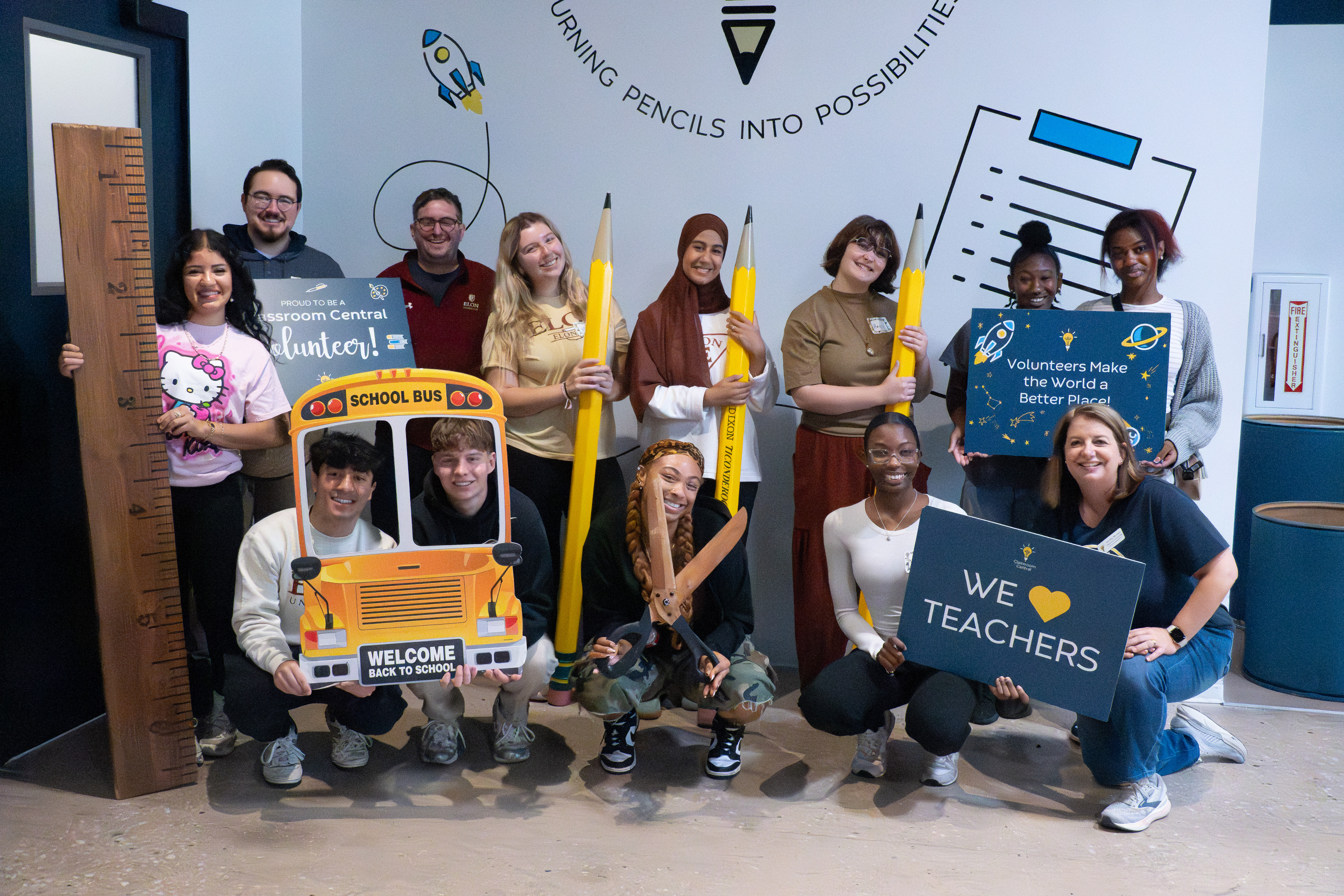 A group of students and staff pose together holding large school-themed props like pencils, scissors, and a ruler during a volunteer event at Classroom Central. Some hold signs that read “Volunteers Make the World a Better Place” and “We Love Teachers.”