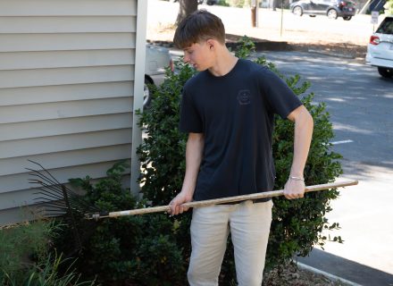 Fritz Graham ’26 uses a rake to clear leaves and debris near a bush outside a building during a community service project. A parking lot and parked cars are visible in the background.