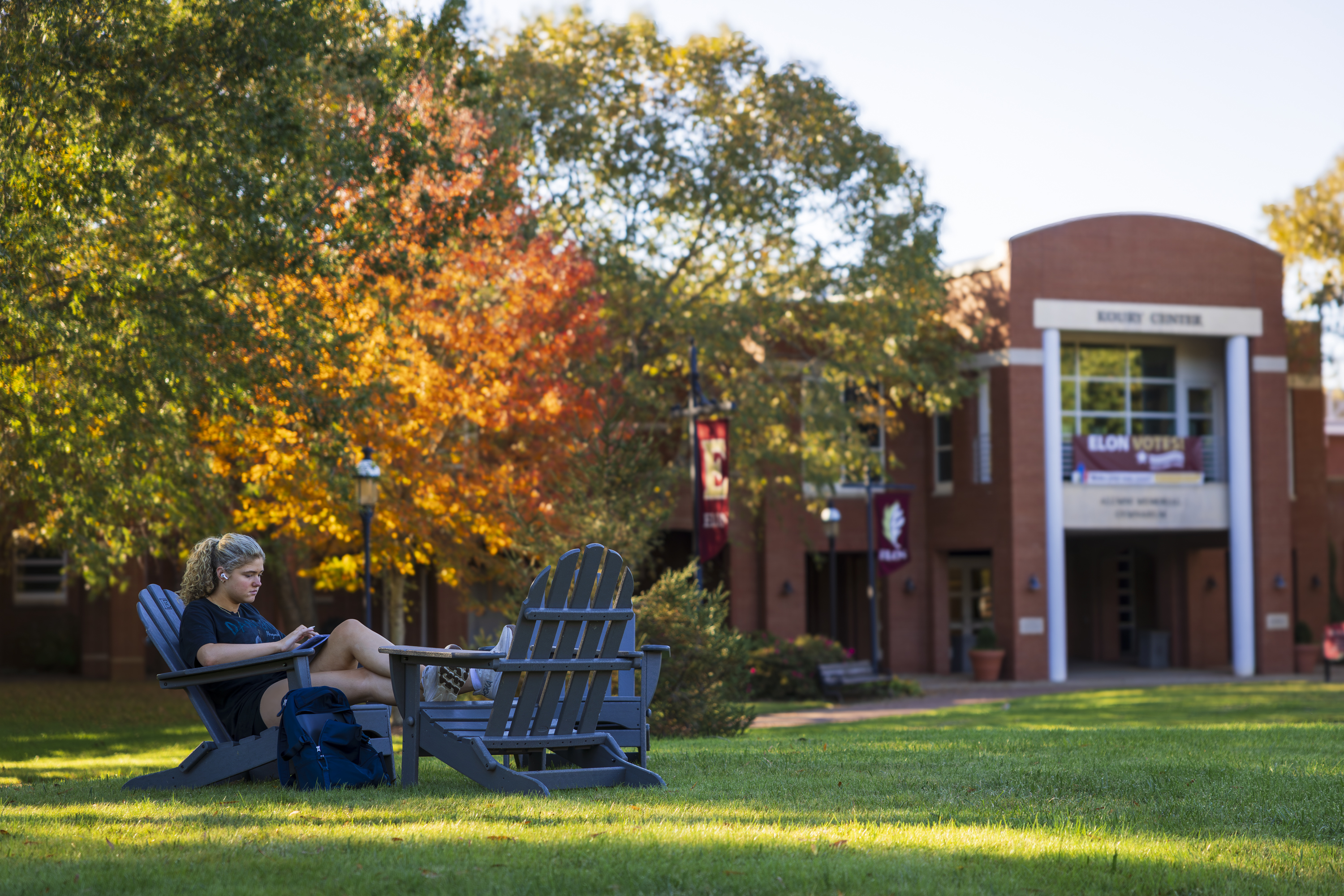 A person is sitting on an Adirondack chair, reading a book on a campus lawn in front of the Moseley Center at Elon University. The surroundings are adorned with colorful autumn leaves.