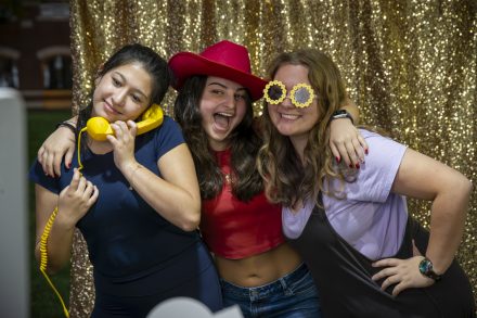 Three students pose playfully in front of a gold sequin backdrop. One holds a yellow phone, another wears a red cowboy hat, and the third has sunflower-shaped glasses.