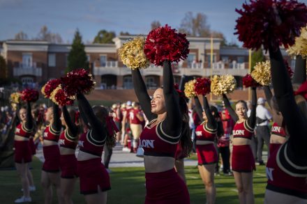 Elon cheerleaders in red and gold uniforms raise pom-poms during a game-day performance on the football field, with fans and players in the background.