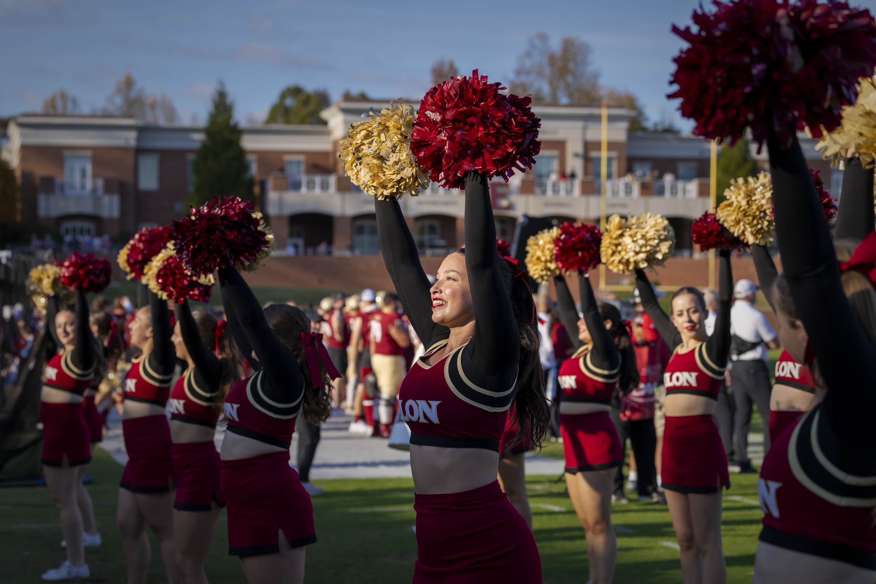 Elon cheerleaders in red and gold uniforms raise pom-poms during a game-day performance on the football field, with fans and players in the background.