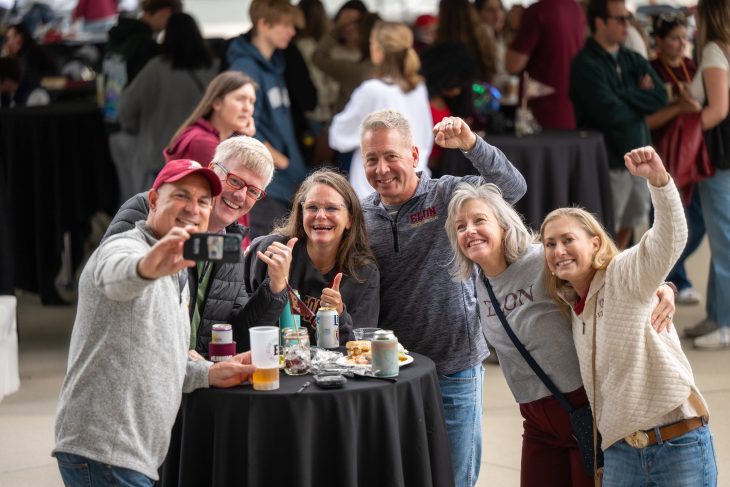 A group of smiling friends at a tailgate take a selfie together at a round table covered with food and drinks.