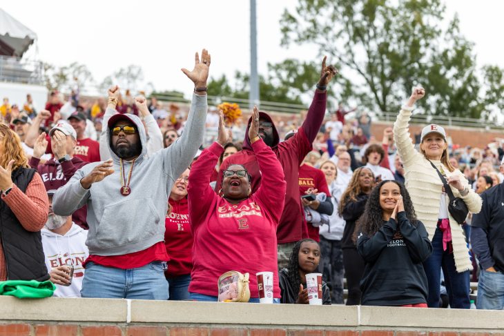 A diverse crowd of Elon fans clap and cheer energetically during a football game.