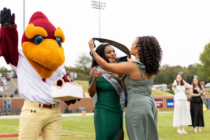 A student in a green dress is crowned Homecoming Royalty on the football field beside the Phoenix mascot.