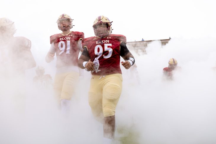 Elon football players in red and gold uniforms emerge dramatically from a cloud of smoke onto the field.