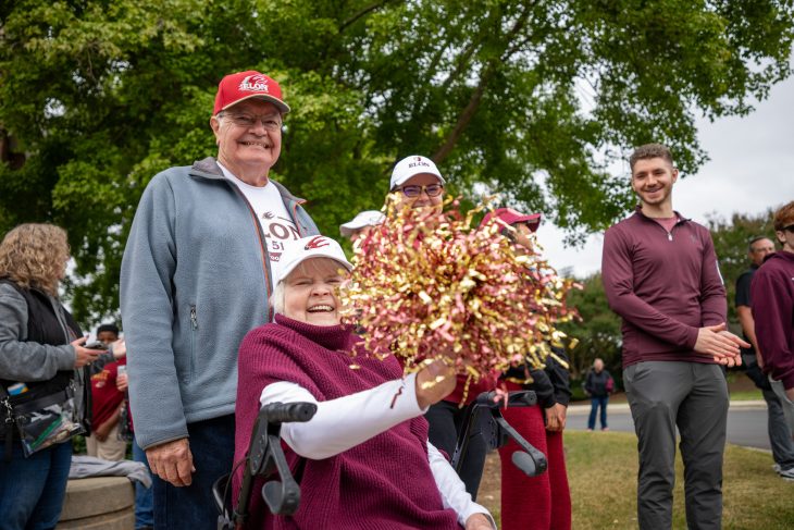 An older couple wearing Elon gear smile proudly, one holding pom-poms while sitting in a wheelchair.