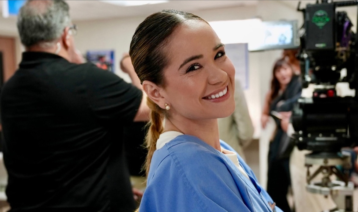 Carmencita Rosales ’20 smiles at the camera wearing light blue scrubs on the set of the Telemundo series “Velvet: Nuevo Imperio.”