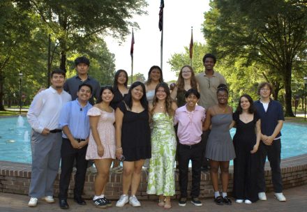 A group of students stand together and smile in front of a fountain on a sunny day, dressed in semi-formal attire.