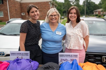 Three women pose for a photo in front of two cars