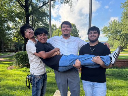 Four students smile outdoors on a sunny day; three stand on a brick walkway holding a fourth student horizontally in their arms.