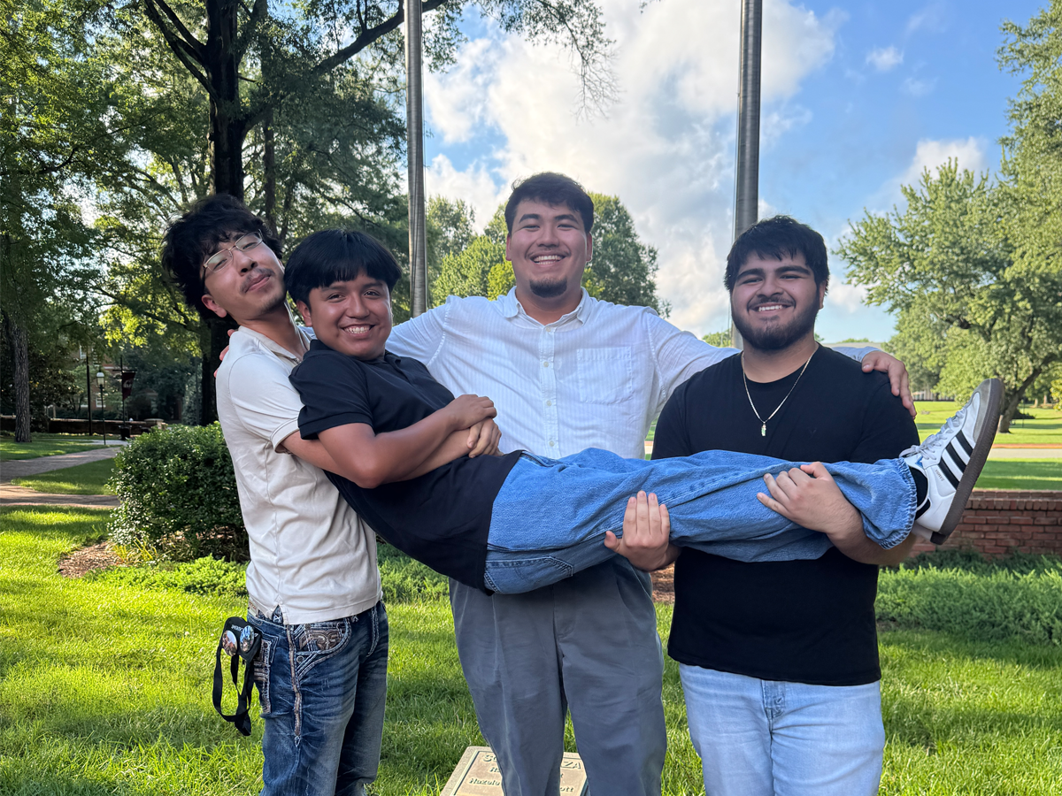 Four students smile outdoors on a sunny day; three stand on a brick walkway holding a fourth student horizontally in their arms.