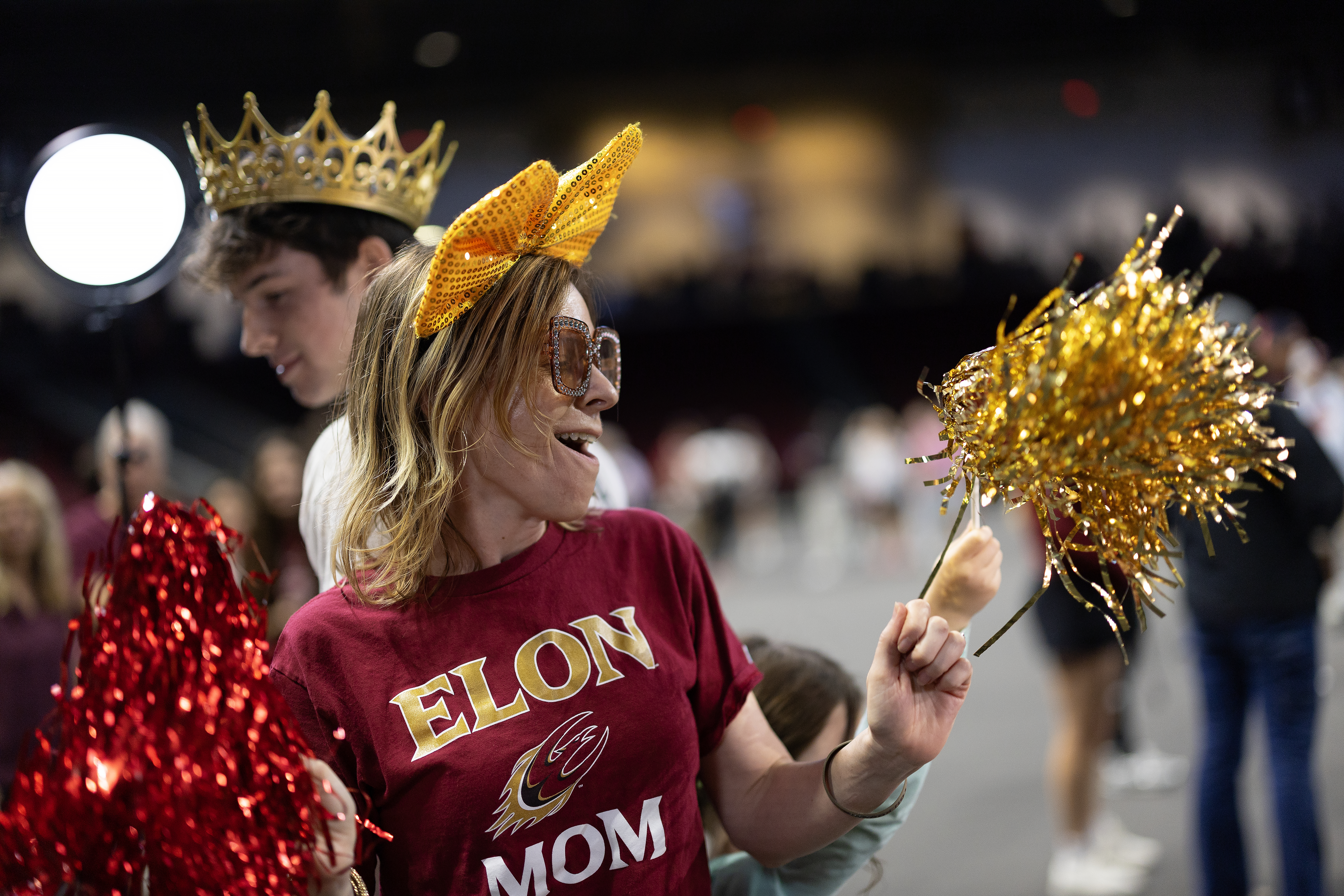 A parent in an “Elon Mom” shirt cheers with pom-poms and festive accessories at Family Weekend.