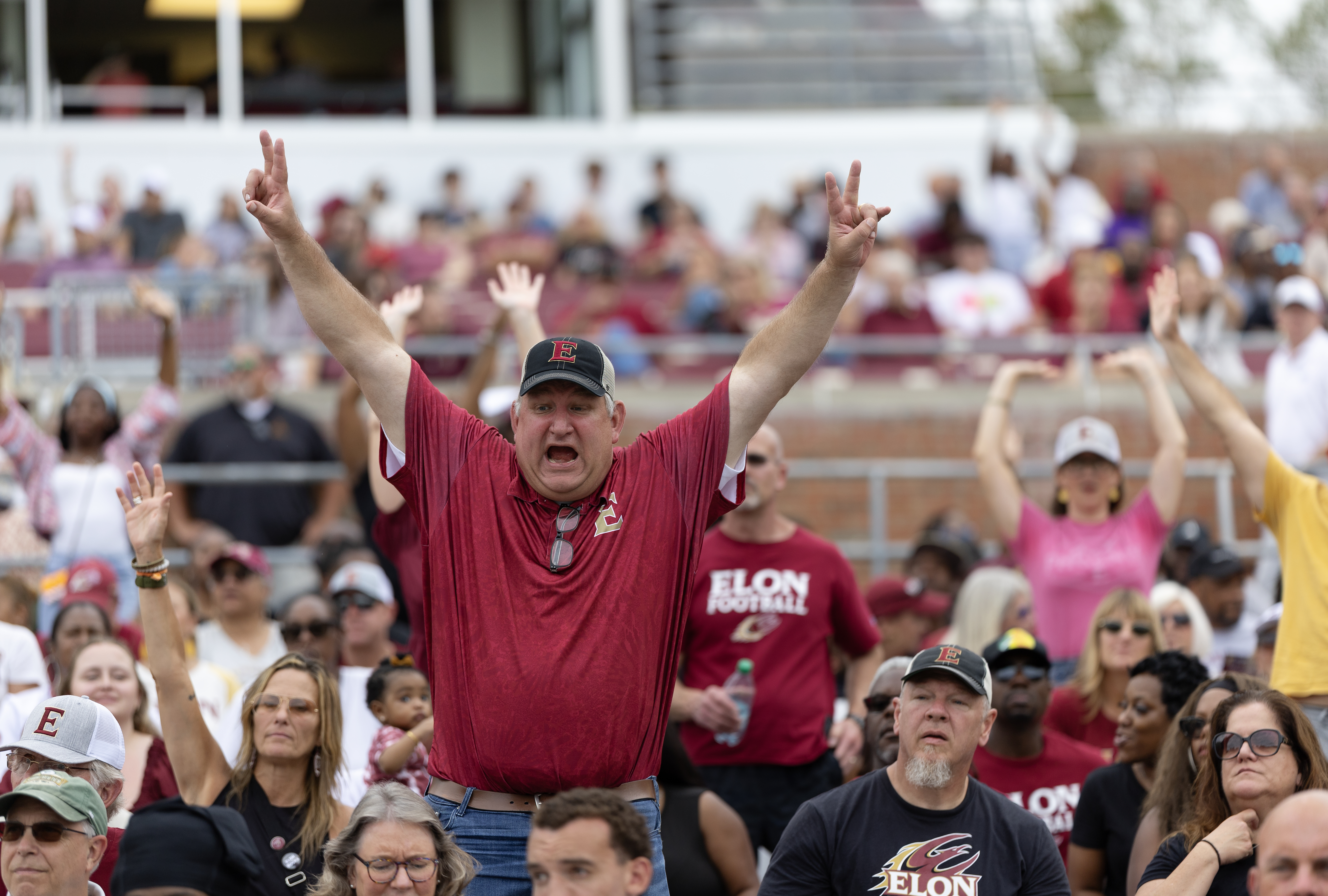 A cheering Elon fan in a red shirt and cap raises both arms with the crowd behind him at a football game.