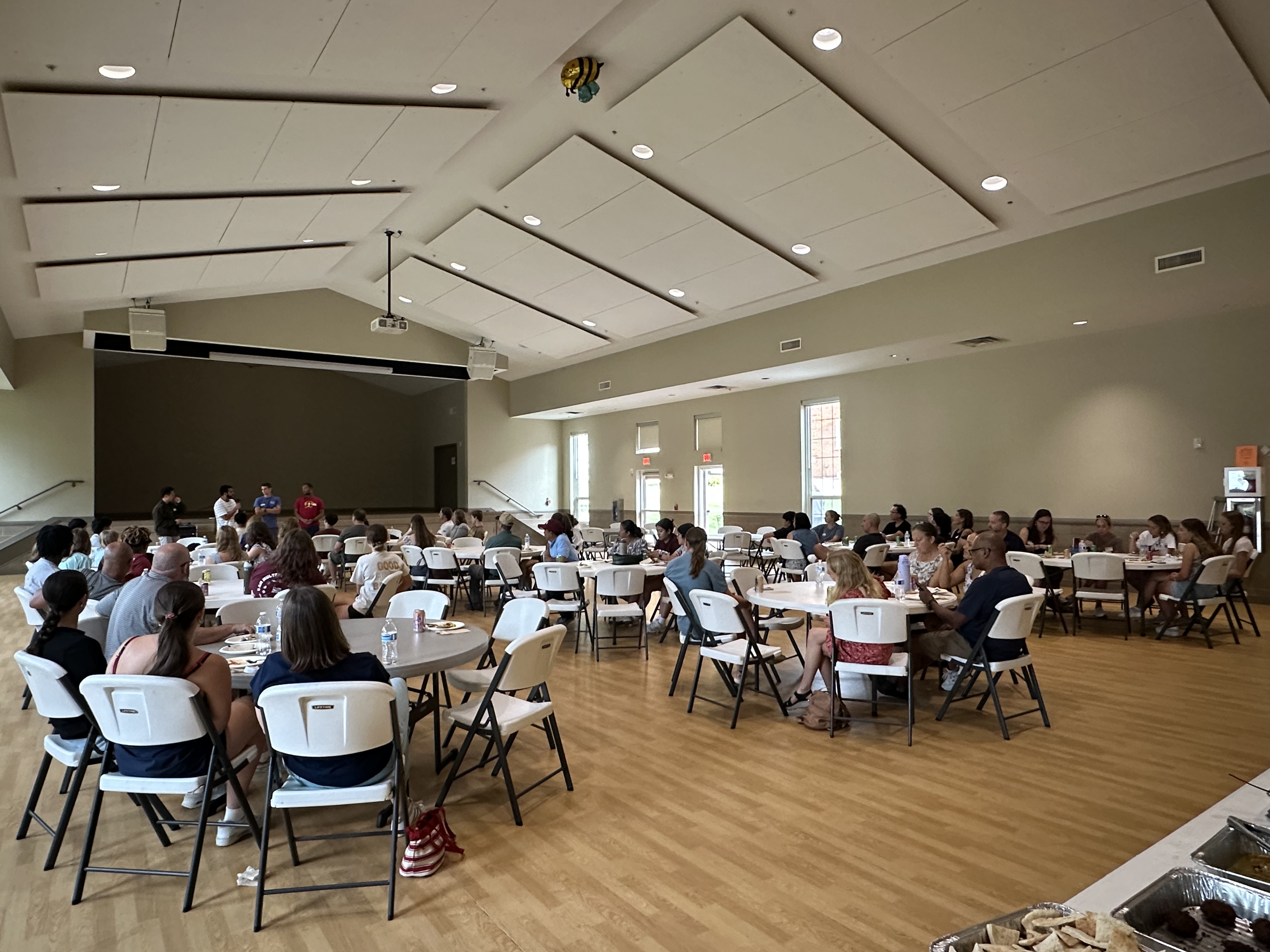 Students and community members sit at round tables in a large hall during a Protestant Life dinner, listening to speakers standing at the front of the room.