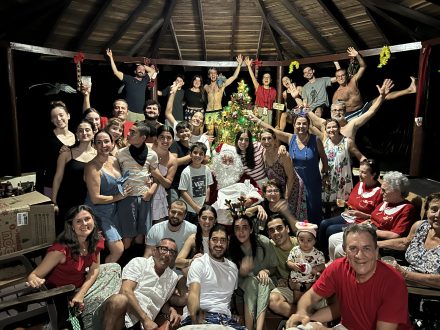 Camila's big family of various ages gather around a decorated Christmas tree under a wooden pavilion, smiling and posing with Santa Claus at the center.