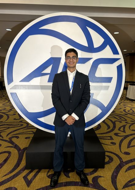 Shaan Gandhi, vestido con un traje negro, se para frente al logo de la Conferencia de la Costa Atlántica y sonríe.
