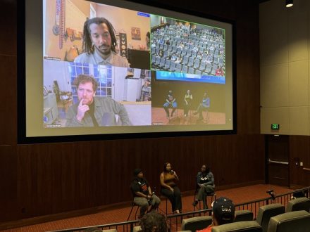 Panelists speak in an auditorium while two virtual participants join via Zoom on a large screen behind them.