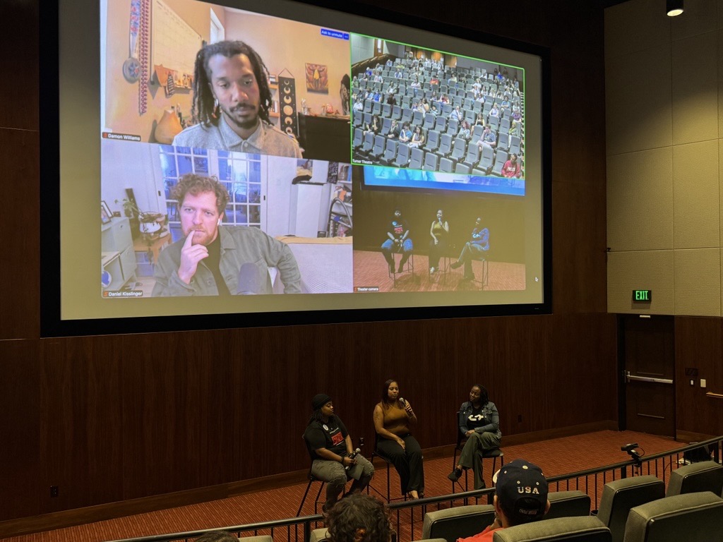 Panelists speak in an auditorium while two virtual participants join via Zoom on a large screen behind them.