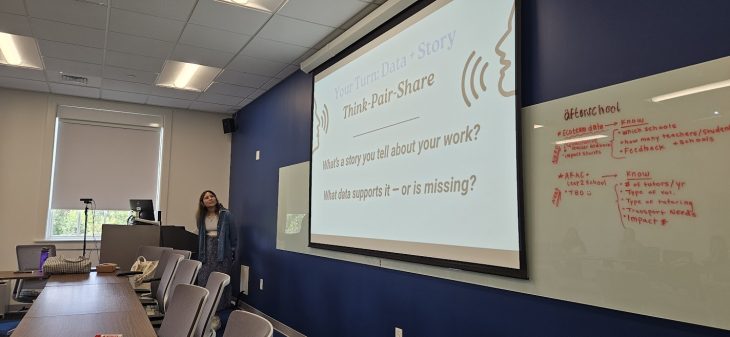 A woman stands in the front of a room facing a presentation screen