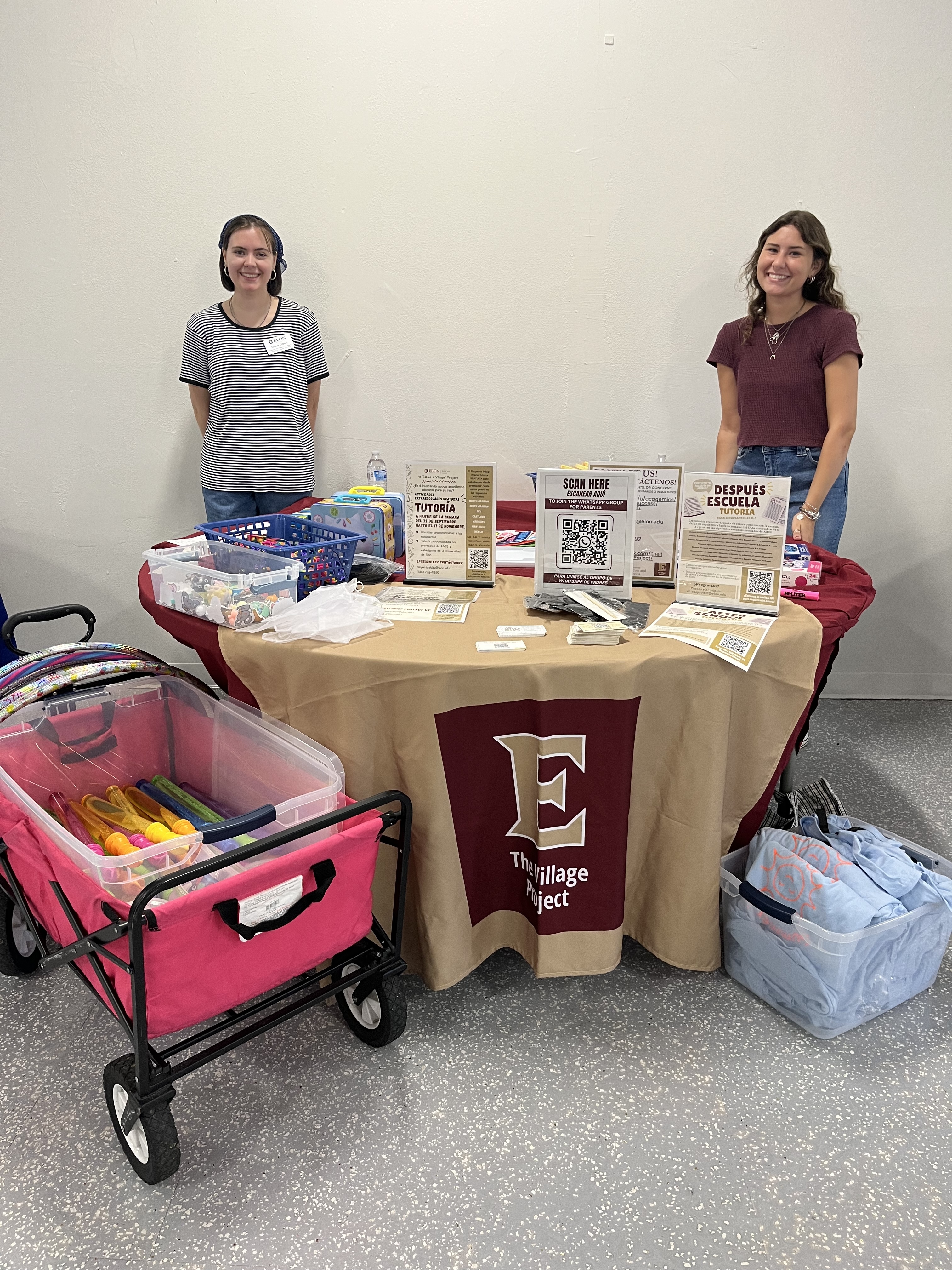 Two women stand in front of a table with items, covered in Elon University banner