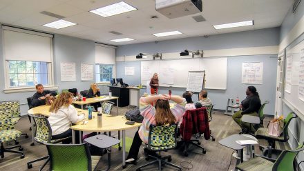 A group of people sit in a classroom and listen to an instructor in front of a white board