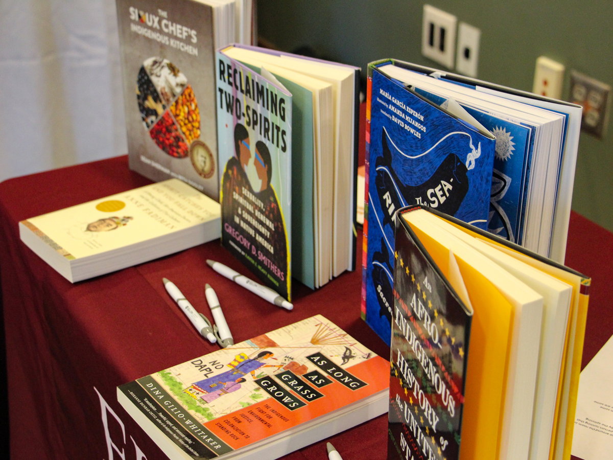 A display of books about Indigenous history and culture arranged on a maroon tablecloth, including titles such as Reclaiming Two-Spirits, The Sioux Chef’s Indigenous Kitchen, and An Afro-Indigenous History of the United States. Several pens are placed beside the books.