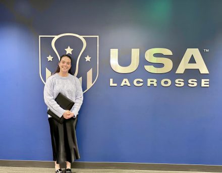 Nyla Lewis stands smiling against a blue wall in front of the USA Lacrosse logo