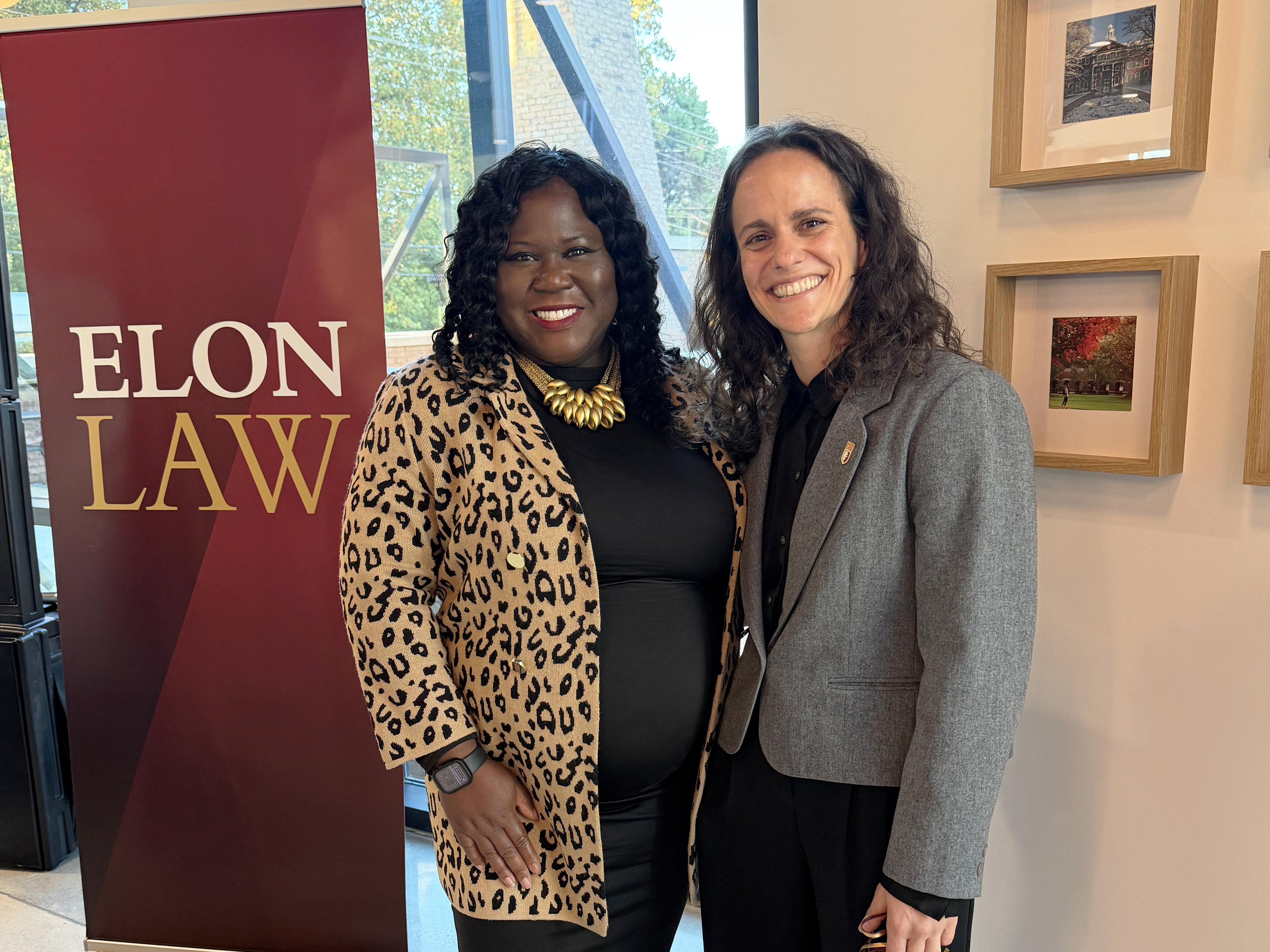 Two women dressed professionally, smiling, in front of an Elon Law banner.