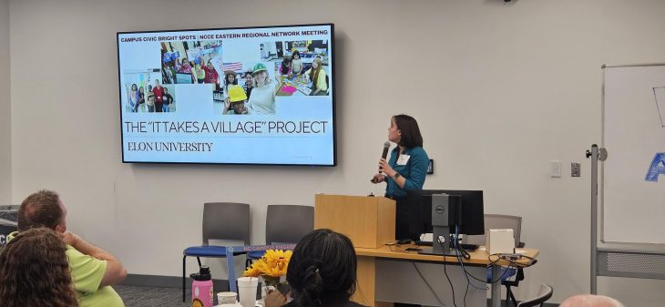 A woman holds a microphone at the front of a room and presents facing a TV that says "The IT Takes A Village" Program Elon University