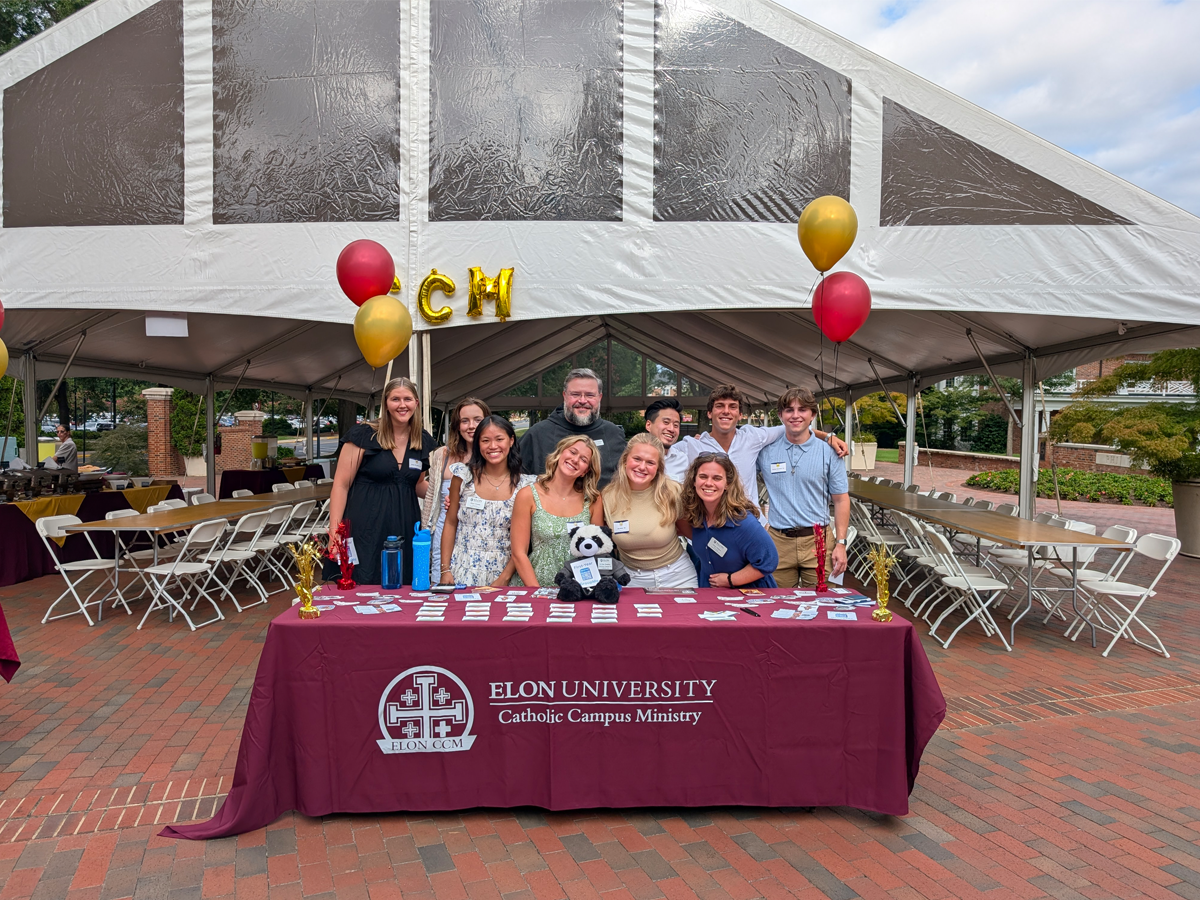 A group of students and staff with Catholic Campus Ministry smile behind a maroon table covered with name tags and balloons at an outdoor welcome event under a large white tent.