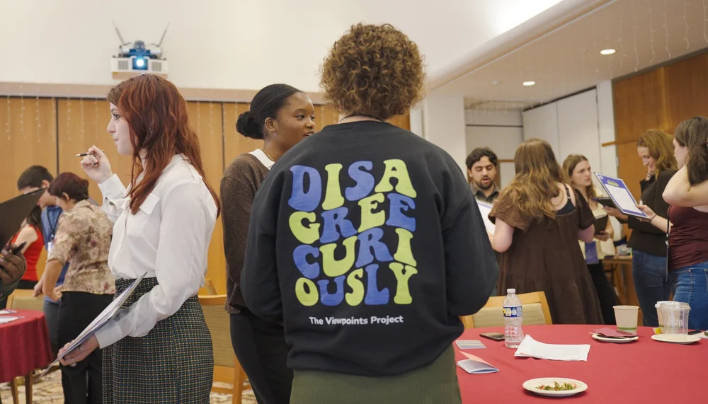 A group of people engage in conversation and note-taking during a workshop or discussion event in a room with tables and papers. The person in the foreground wears a black sweatshirt that reads “DISAGREE CURIOUSLY – The Viewpoints Project.”