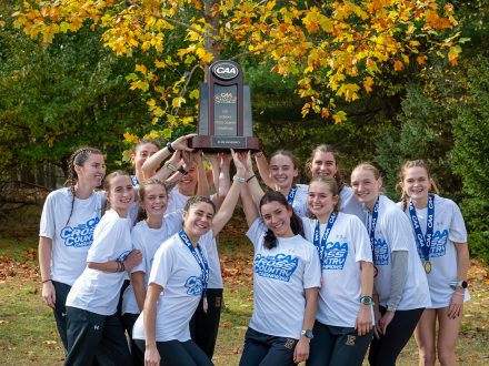 Elon womens cross country team stand in a circle in white shirts and hold up a trophy