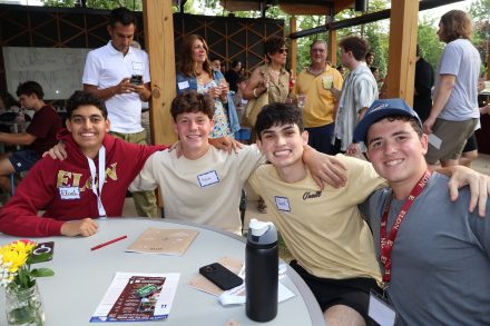 : Four students sit together at a round table during the Jewish Life welcome dinner, smiling with arms around each other. The table holds papers, a water bottle, and a small vase of flowers.