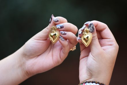 Two hands hold golden earrings in the shape of hearts. Her nail polish is an earthy brown white swirls.
