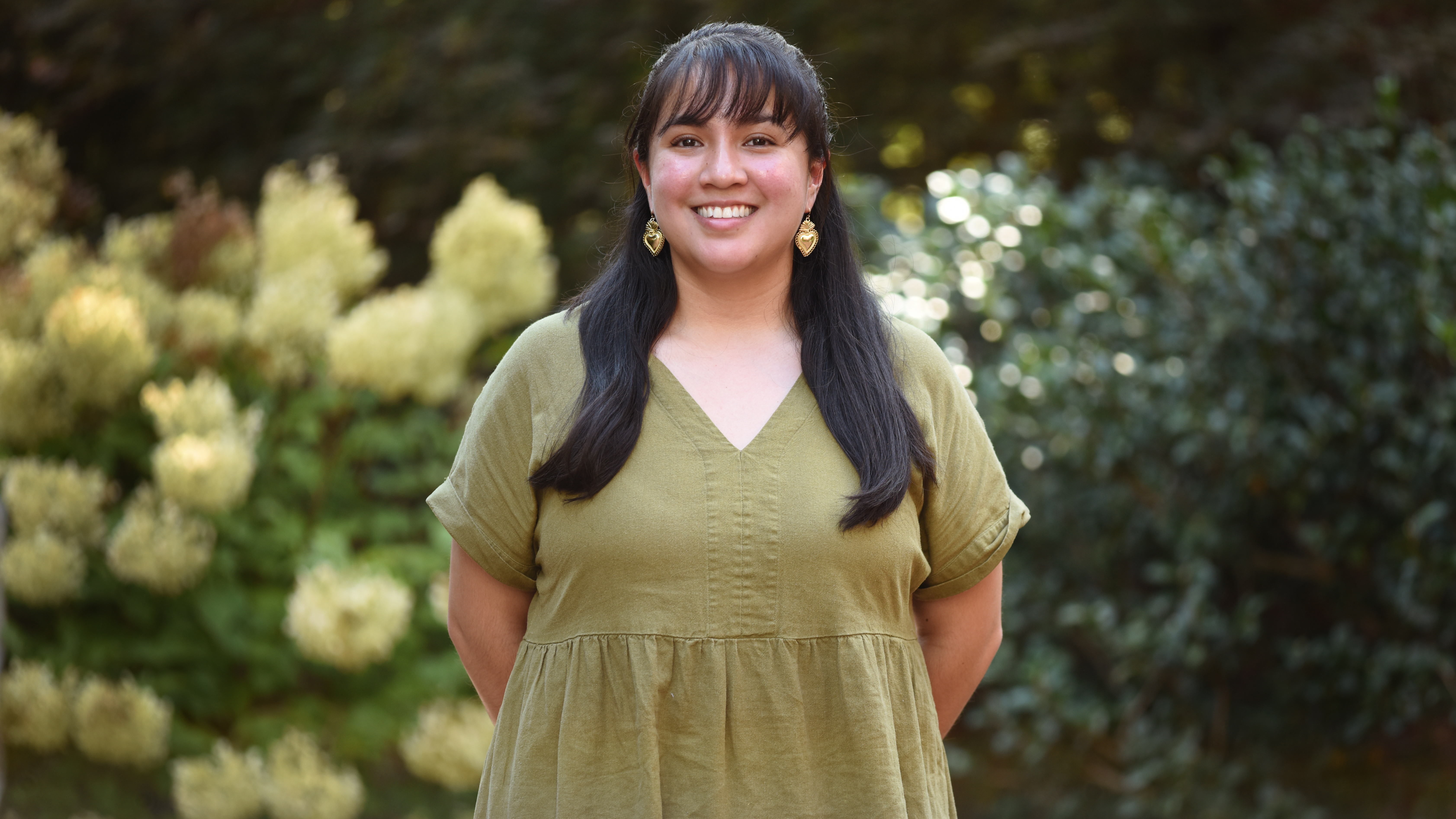 A woman with long dark hair, earrings and an olive green dress stands in front of greenery