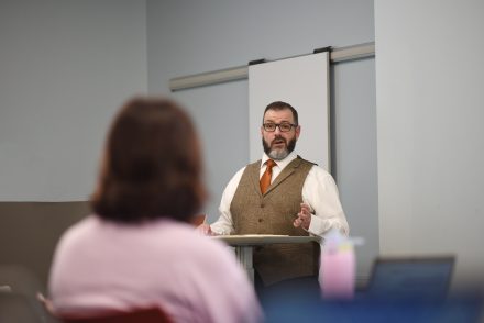 A man with short dark brown hair, glasses and a gray and black bear stands in front of a podium addressing an audience member. He is wearing a white button shirt with a brown vest and red tie.