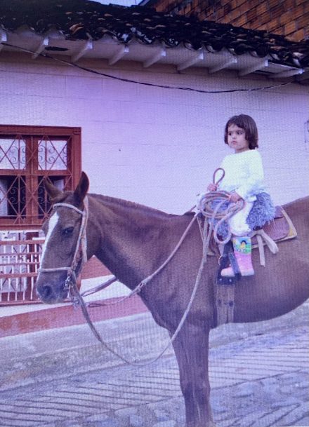 Camila as a child on a brown horse holding a rope, with a traditional white building in the background.