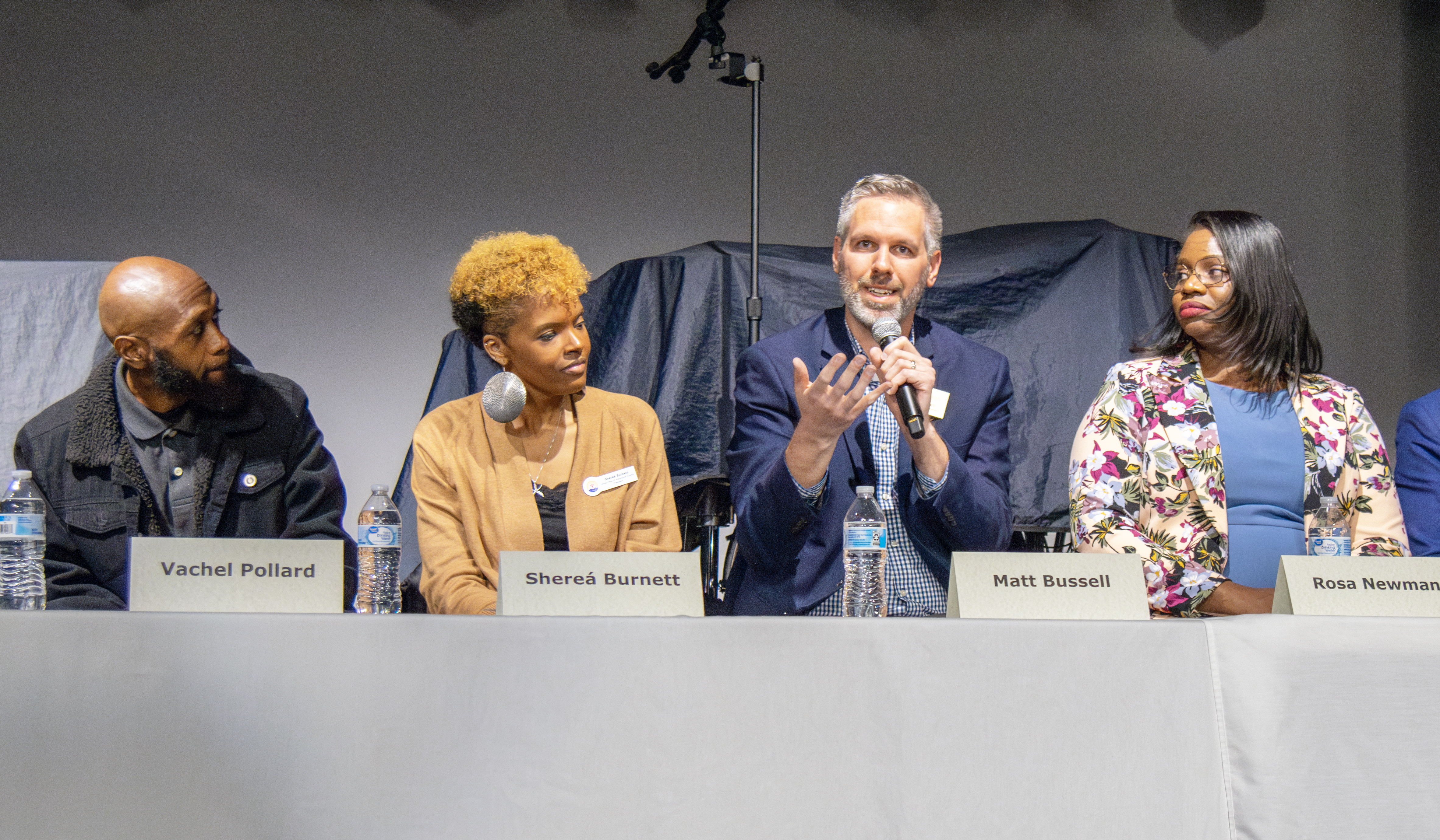 A man in a blue blazer gestures while speaking in a microphone. Three seated around him at a table listen.
