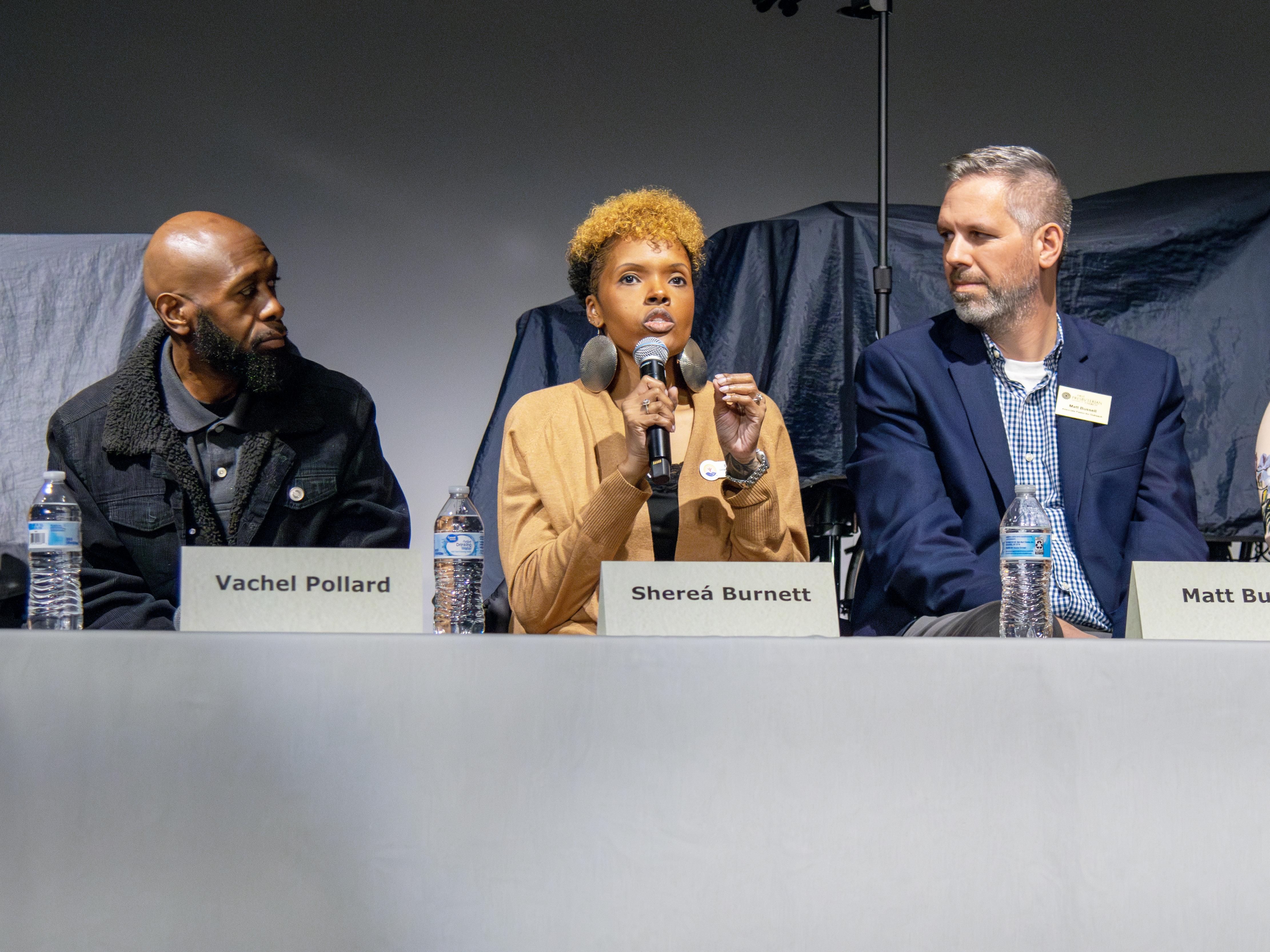 A woman in a tan suit speaks into a microphone and gestures to make a point. Two panelists face her, listening.