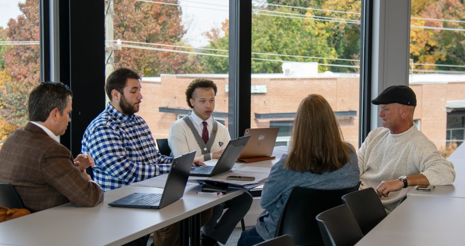 A group of five people around a long table in a large, windowed classroom. They are conversing and reviewing documents