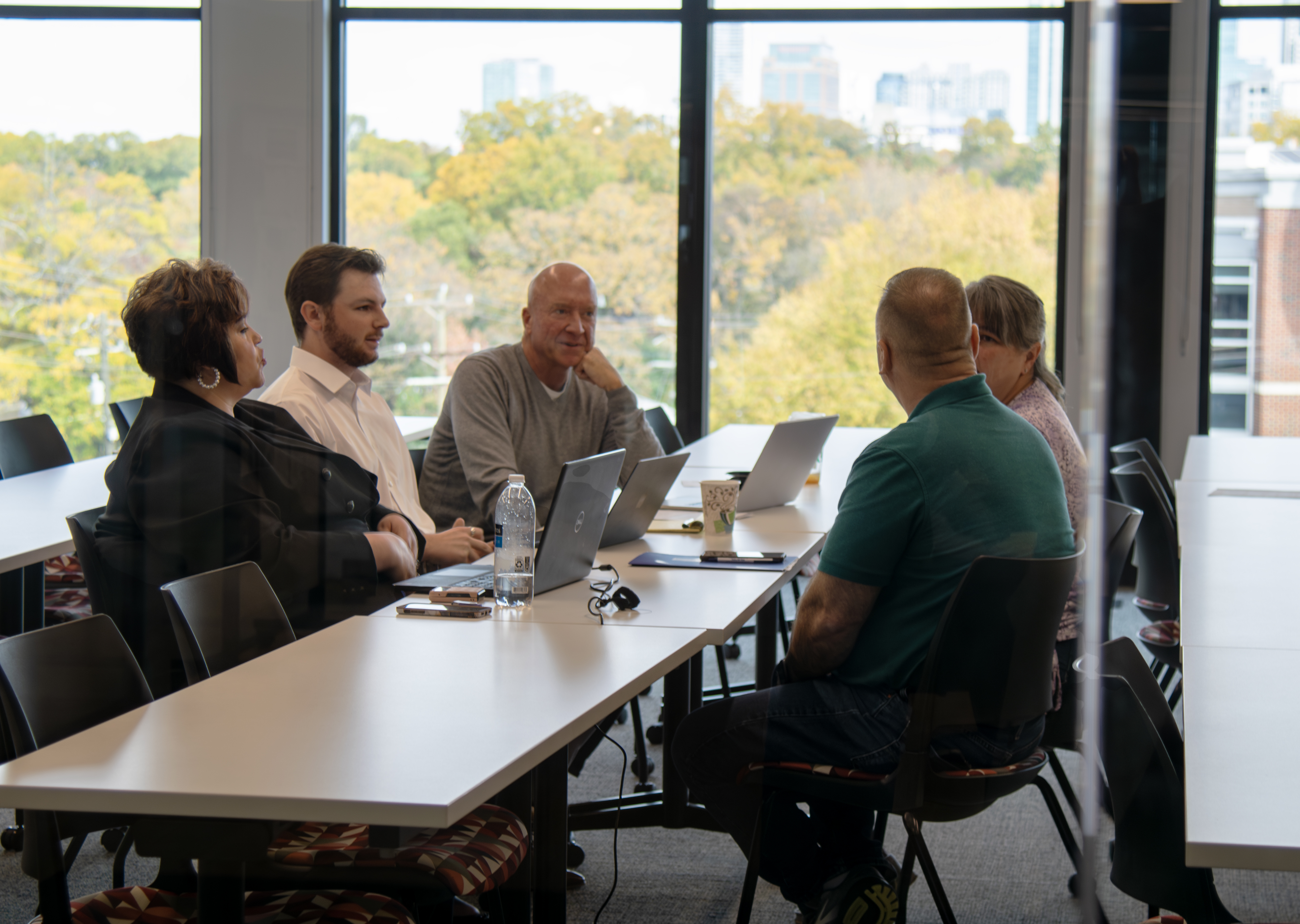 A group of five people around a long table in a large, windowed classroom with the Charlotte skyline behind them.