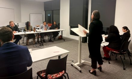 A woman show from behind, speaking at a podium.