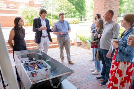 A group of people stand around an engineering project. Three people are speaking to four people