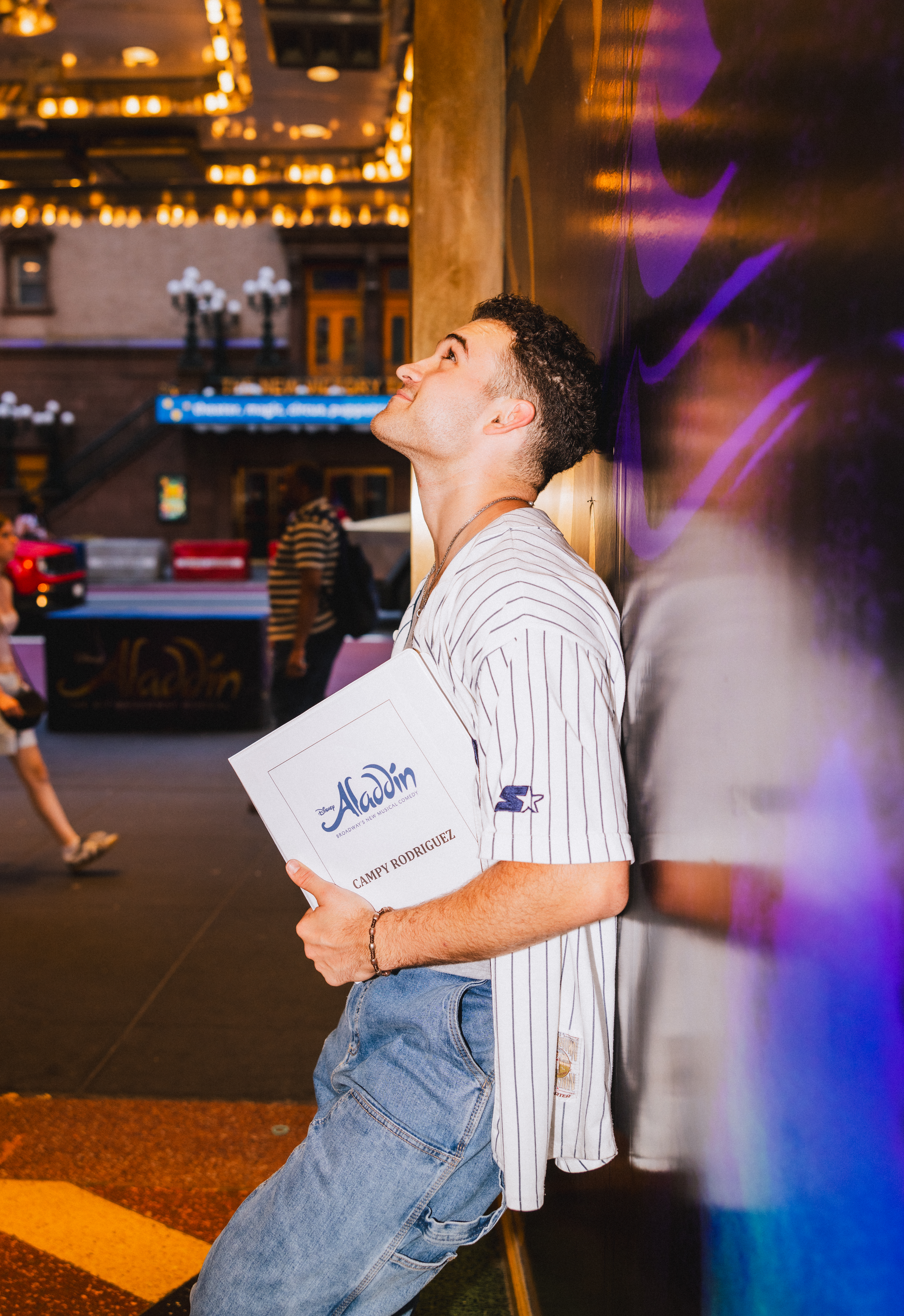 A man leans against a wall holding a binder that says "Aladdin" on the front