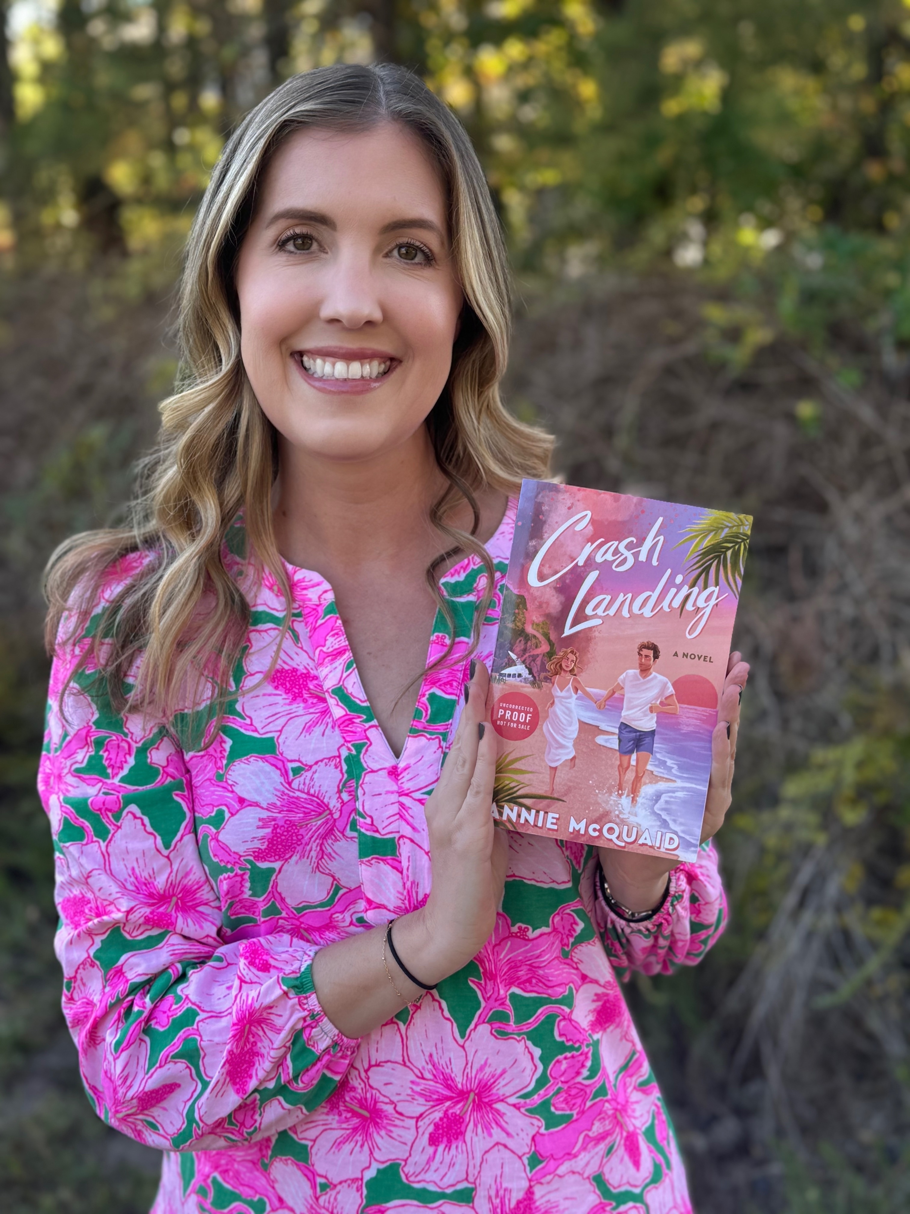 A woman holds up the book "Crash Landing". She is wearing a bright pink dress.
