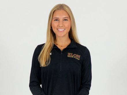 A woman in an Elon track and field quarter zip poses for a photo in front of a white background