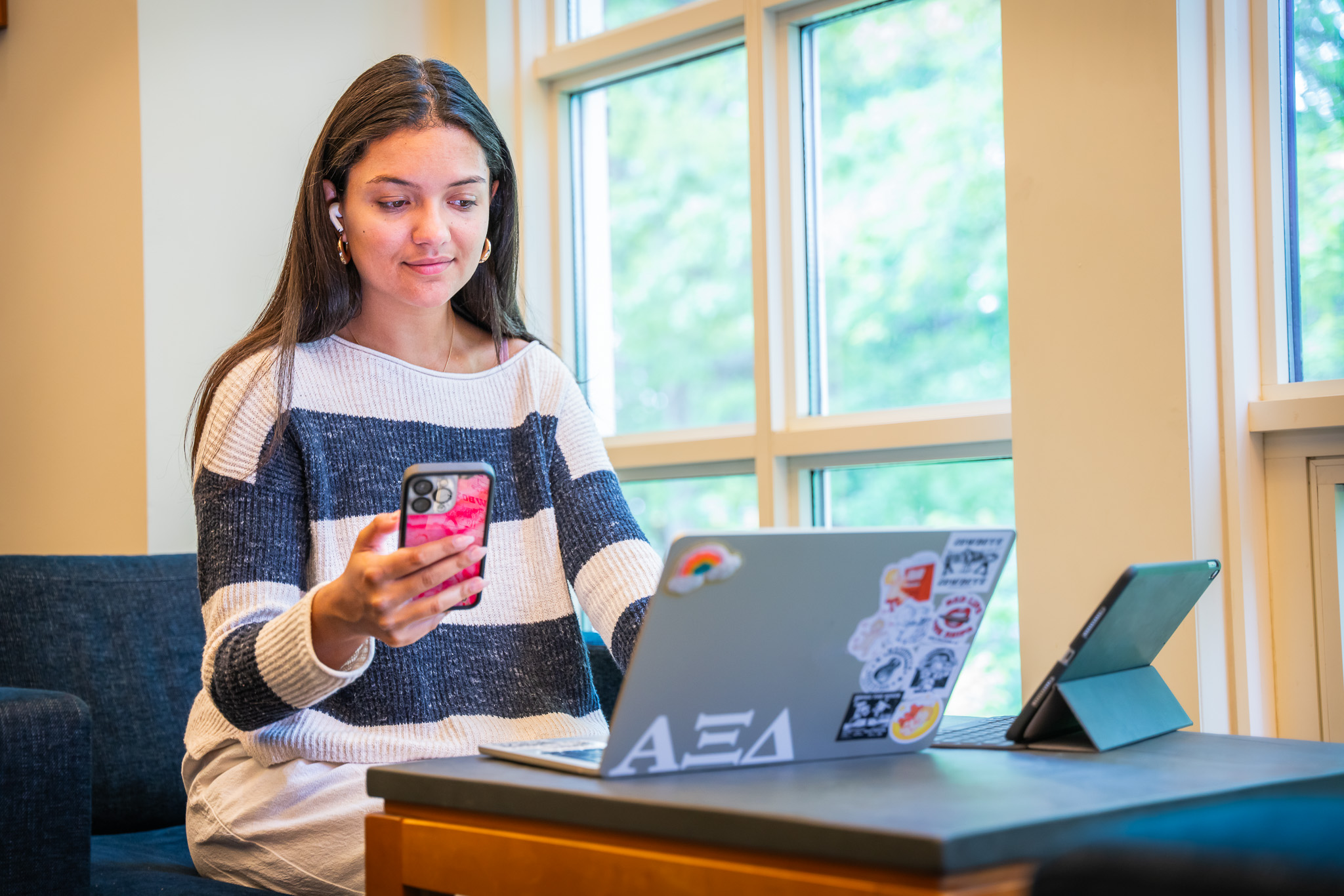 A student with long dark hair sits in a bright study area by large windows. She wears a cream and navy striped sweater and white pants, and is seated at a small table with a laptop covered in stickers, a tablet in a case, and a smartphone in her hand. Soft natural light fills the space, and trees are visible through the windows behind her.