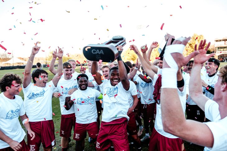 Elon soccer players celebrate a win while cheering together. One player in the middle holds up a trophy